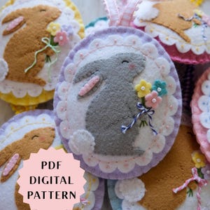 Könnte beinhalten: Handgemachte Osterhasen-Ornamente aus Filz. Jedes Ornament zeigt einen Hasen mit Blumen, mit einem gewellten Rand und einem Band zum Aufhängen. Die Ornamente sind in verschiedenen Farben erhältlich, darunter Braun, Grau und Weiß, mit Pastellakzenten.