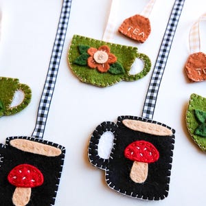 May include: Felt tea bag and teacup ornaments in green and black, with floral and mushroom designs. Each ornament has a stitched border and a ribbon for hanging. The tea bag tags are brown with "TEA" stitched on them.