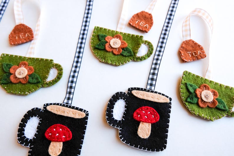 May include: Felt tea bag and teacup ornaments in green and black, with floral and mushroom designs. Each ornament has a stitched border and a ribbon for hanging. The tea bag tags are brown with "TEA" stitched on them.