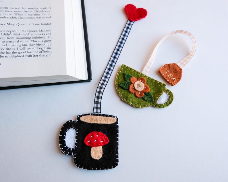 May include: Handmade felt bookmarks featuring a black mug with a mushroom design and a green teacup with a flower. The bookmarks have a gingham ribbon and a red heart. The teacup bookmark has the word "TEA" on it.