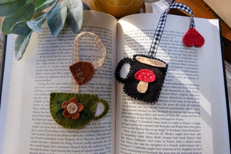 May include: Three handmade felt bookmarks rest on an open book. One is a tea bag shape with "TEA" text, another is a mug with a mushroom, and the last is a heart. The bookmarks are on a book with text.
