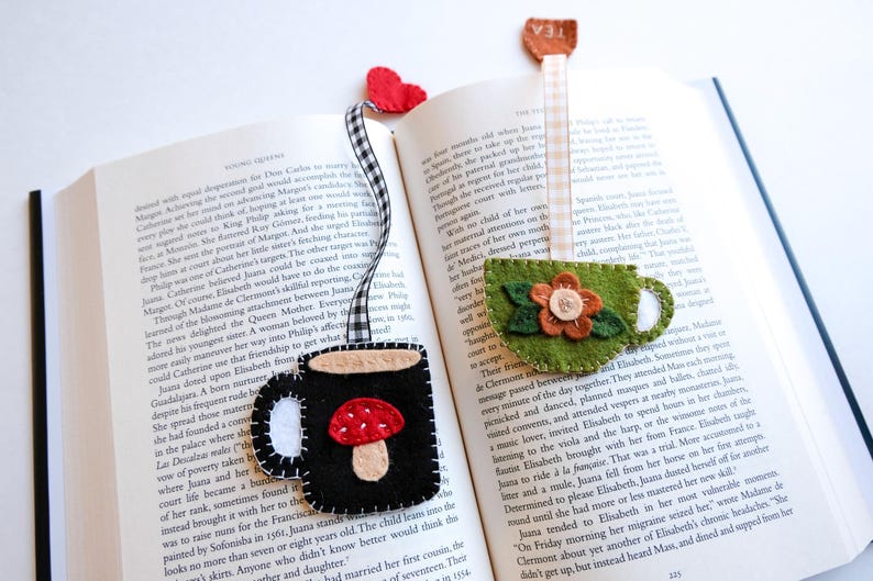 May include: Handmade felt bookmarks with whimsical designs. One features a black mug with a mushroom, another a green teacup with a flower, and a third with a heart. They are placed in an open book.