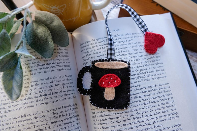 May include: A handmade black felt bookmark shaped like a mug with a red mushroom design. The bookmark has a black and white checkered ribbon and a red heart. The bookmark is placed on an open book.