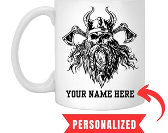 Puede incluir: Taza de cerámica blanca con una ilustración en blanco y negro de un guerrero vikingo con hachas. El texto "YOUR NAME HERE" está impreso debajo de la ilustración.