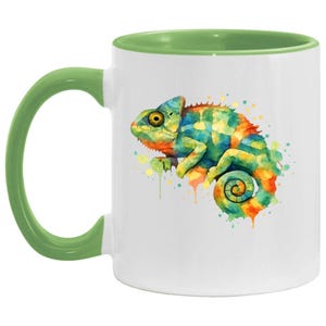 May include: A white ceramic mug featuring a vibrant watercolour illustration of a chameleon. The mug has a light green handle and a matching light green interior rim. The chameleon design displays a mix of blue, yellow, and orange paint splatters.