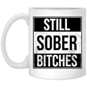 May include: White ceramic mug with a black and white graphic. The text on the mug reads "STILL SOBER BITCHES" in a bold, distressed font. The mug has a curved handle and a glossy finish.