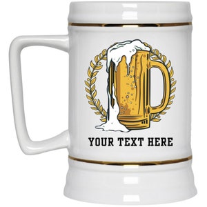 Könnte beinhalten: Ein weißer Keramik-Bierkrug mit goldfarbenem Rand und einer Grafik eines Bierkruges mit überlaufendem Schaum. Die Grafik ist von einem Kranz aus Weizen umgeben. Der Text "YOUR TEXT HERE" ist unter der Grafik gedruckt.