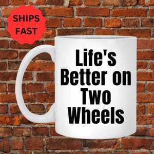 以下が含まれることがあります： 白いセラミック製のコーヒーカップで、黒い文字で「Life's Better on Two Wheels」と書かれています。