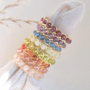 May include: A stack of colorful beaded rings. Each ring features a gold-colored wire frame holding a series of small, round gemstones in various colors, including pink, orange, green, white, blue, purple, and red. The rings are resting on a white cloth.