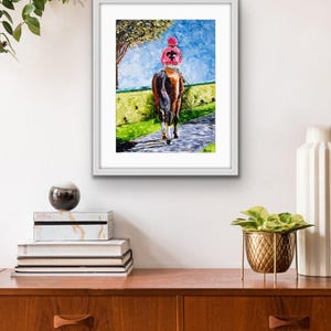 May include: Framed artwork depicting a horse and jockey on a racetrack. The jockey wears a pink cap and jacket with a black design. The horse is brown, and the background features a blue sky, green grass, and a tree.
