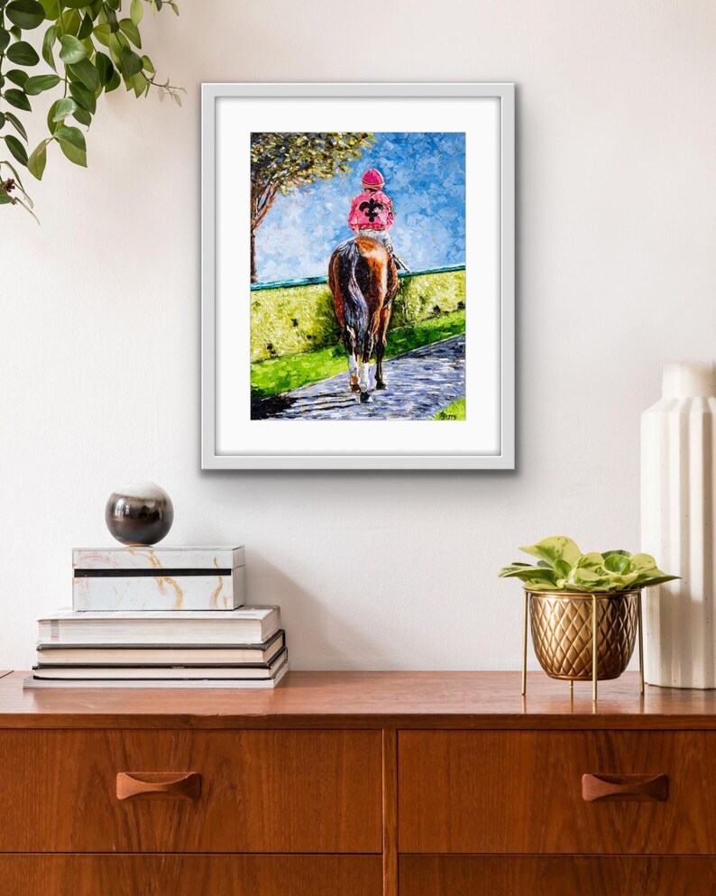 May include: Framed artwork depicting a horse and jockey on a racetrack. The jockey wears a pink cap and jacket with a black design. The horse is brown, and the background features a blue sky, green grass, and a tree.