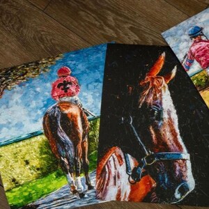 May include: A collection of three equestrian-themed art prints. One print depicts a racehorse and jockey in red and black, another shows a close-up of a horse's head, and the third features a jockey in pink and blue. The artworks are displayed on a wooden surface.