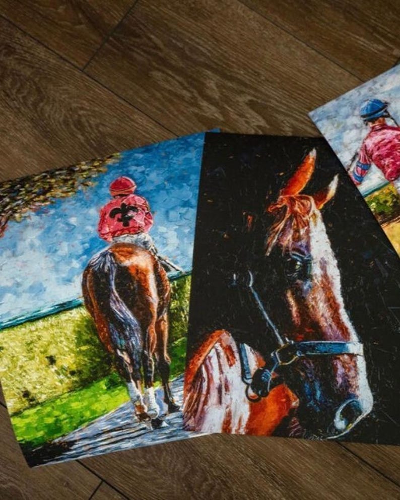 May include: A collection of three equestrian-themed art prints. One print depicts a racehorse and jockey in red and black, another shows a close-up of a horse's head, and the third features a jockey in pink and blue. The artworks are displayed on a wooden surface.