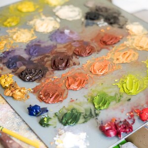 May include: A close-up of a painter's palette with various colors of oil paint. The palette is filled with blobs of yellow, orange, red, green, blue, and brown paint. A yellow pencil and a palette knife are in the foreground.
