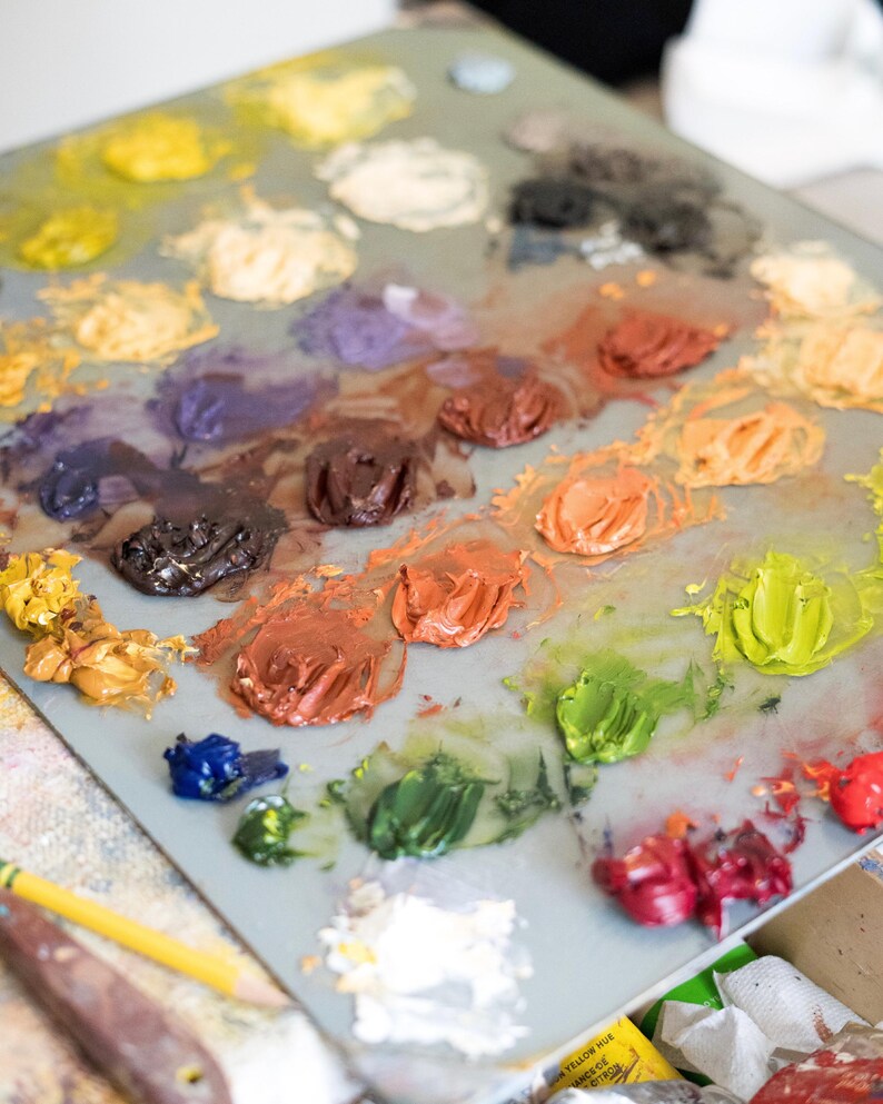 May include: A close-up of a painter's palette with various colors of oil paint. The palette is filled with blobs of yellow, orange, red, green, blue, and brown paint. A yellow pencil and a palette knife are in the foreground.