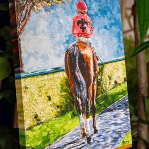 May include: Oil painting on canvas of a jockey on a horse, viewed from behind. The jockey wears a red cap and jacket with a black fleur-de-lis. The horse is brown and white, and the background features a blue sky, green field, and a tree.