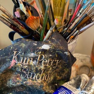 May include: A collection of paintbrushes of various sizes and shapes, some with colorful paint on the bristles, are displayed in a metal container and a paint-splattered apron. The apron has the text "Kim Petry Studio" embroidered on it. Two tubes of Winsor & Newton oil paint are visible.