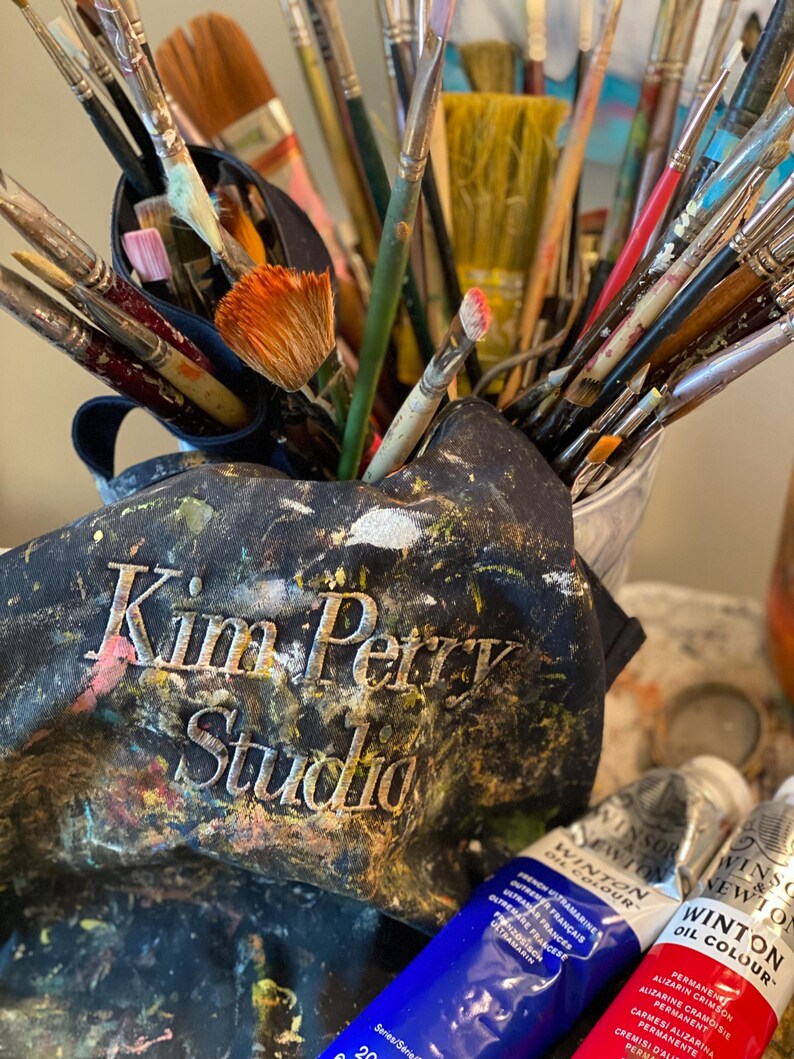 May include: A collection of paintbrushes of various sizes and shapes, some with colorful paint on the bristles, are displayed in a metal container and a paint-splattered apron. The apron has the text "Kim Petry Studio" embroidered on it. Two tubes of Winsor & Newton oil paint are visible.
