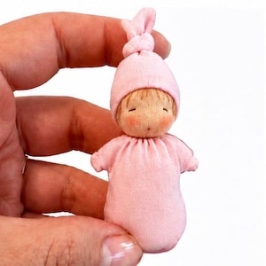 May include: A miniature doll with a light skin tone, rosy cheeks, and closed eyes. The doll wears a soft pink fabric outfit that covers the body and a matching pink hat with a small knot at the top. The doll is small enough to be held in one hand.