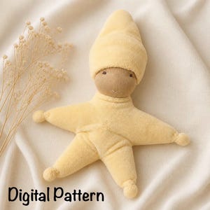 May include: A soft, pale yellow star-shaped doll with a matching pointed cap, featuring simple embroidered eyes and a small mouth. The doll has a plush, textured surface and is positioned next to a sprig of dried baby's breath flowers.