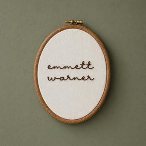 May include: An oval embroidery hoop featuring the names "emmett warner" stitched in brown thread on white fabric. The wooden hoop has a brass clasp. The background is a solid olive green.