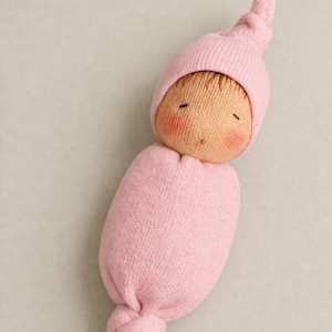 以下が含まれることがあります： 柔らかいピンク色の布に包まれた手作りの赤ちゃん人形。人形は、ニットのピンクの帽子、目を閉じた丸い顔、バラ色の頬をしています。人形の体は、下で結ばれたお揃いのピンク色の布で包まれています。