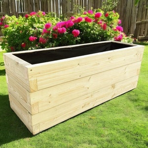 May include: A wooden planter box with a black liner. The planter is made of natural wood and has a rectangular shape. It is sitting on a green lawn.