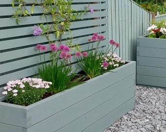 May include: Three rectangular planters made of light gray wood with a slatted design. The planters are filled with various plants and flowers, including pink and purple flowers. The planters are sitting on a bed of gray gravel.