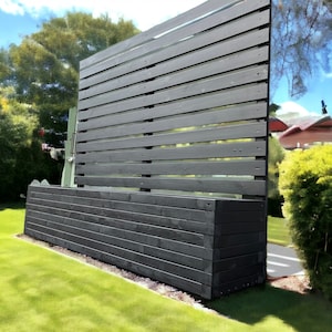 May include: A black wooden planter box with a black wooden slatted fence behind it. The planter box is sitting on a green lawn.