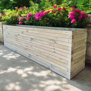 May include: A long, rectangular wooden planter box with a natural finish. The planter is made of horizontal planks and is sitting on a concrete surface.