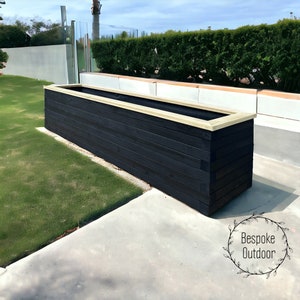 May include: A long, rectangular wooden planter box with a black stain finish and a light brown top trim. The planter is sitting on a concrete patio with green grass in the background.