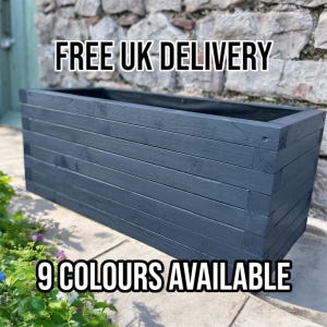 May include: A rectangular, dark blue wooden planter box with horizontal slats. The planter is empty and sits on a stone surface, with greenery and flowers in the foreground. The text "9 COLOURS AVAILABLE" is at the bottom.