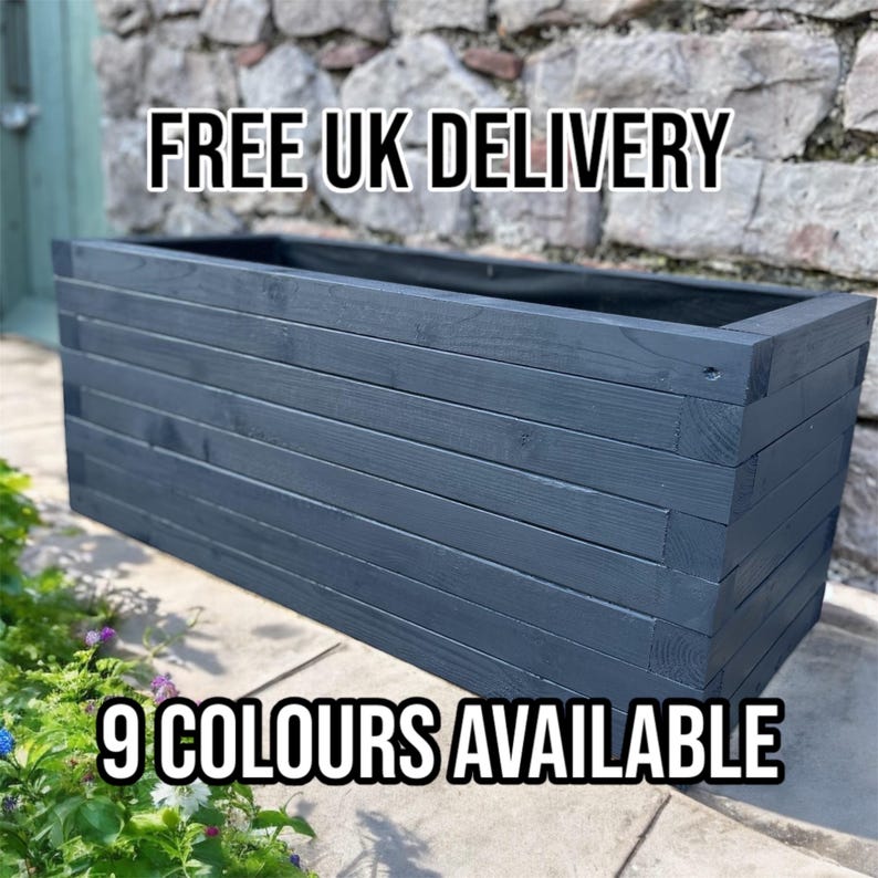 May include: A rectangular, dark blue wooden planter box with horizontal slats. The planter is empty and sits on a stone surface, with greenery and flowers in the foreground. The text "9 COLOURS AVAILABLE" is at the bottom.