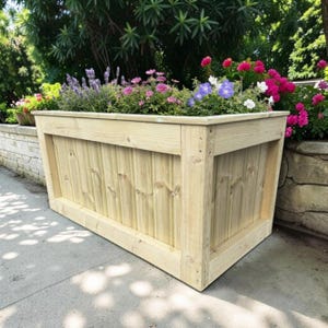 May include: A rectangular wooden planter box with a light natural finish, featuring vertical paneling and thick corner posts. The planter is filled with a variety of blooming flowers, including purple lavender, pink blossoms, and blue petals.