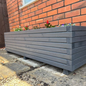 May include: A long, rectangular planter made of dark grey wood with horizontal slats. The planter is sitting on a grey concrete patio and has a few small plants growing in it.