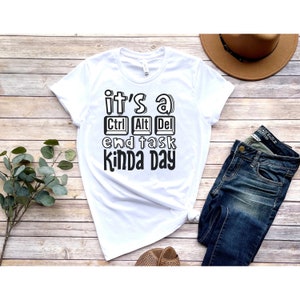 May include: White t-shirt with black text that reads "it's a Ctrl Alt Del end task kinda day". The shirt is laid out on a wooden surface with a pair of brown sandals, a pair of blue jeans, and a brown hat.