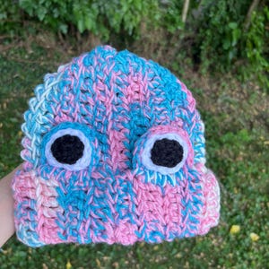Op de afbeelding: Een handgemaakte gehaakte beanie in roze, blauw en wit. De hoed heeft twee grote, ronde, zwart-witte oogapplicaties. De beanie is ontworpen met een gestructureerd steekpatroon.