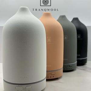 May include: Four ceramic essential oil diffusers in white, light orange, gray, and black. The diffusers are all the same shape and have a round base with a dome-shaped top. The diffusers have a label that says "PISA" and "TRANQWOOL".