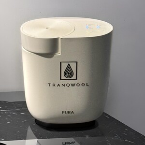 May include: A white cylindrical diffuser with a black base. The diffuser has a black logo that says "TRANQWOOL" and "PURA" on the front.