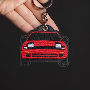 May include: Red embroidered car keychain with black details. The keychain is shaped like the front of a car with headlights and a grill.