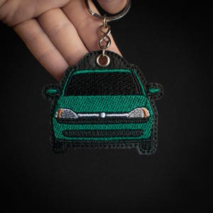 以下が含まれることがあります： 黒のディテールが付いた緑の車のキーホルダー。キーホルダーは革製で、銀色の金属製のリングが付いています。