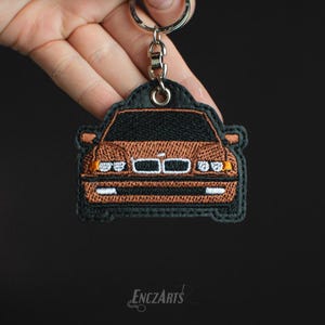 Może przedstawiać: Brązowy, haftowany brelok do kluczy w kształcie samochodu na czarnym tle. Samochód to BMW z srebrną kratką i białymi reflektorami. Brelok jest przymocowany do srebrnego kółka na klucze.