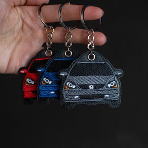 Op de afbeelding: Drie geborduurde autosleutelhanger, elk met een andere kleur: rood, blauw en grijs. De sleutelhangers zijn bevestigd aan zilveren sleutelringen.