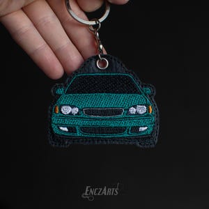 May include: A teal green car keyring with black stitching and a silver key ring. The keyring is embroidered with the front of a car.