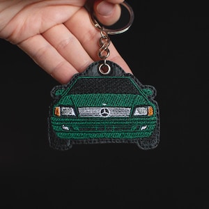 Może przedstawiać: Zielony brelok do kluczy w kształcie samochodu z srebrnym k&oacute;łkiem. Samoch&oacute;d jest wyszyty białymi reflektorami i srebrnym logo Mercedes-Benz.