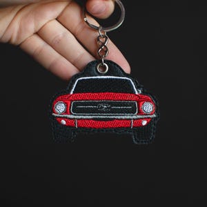 Peut inclure: Un porte-clés brodé en forme de voiture rouge et noir avec un anneau argenté. La voiture présente un avant rouge avec des détails blancs, un toit noir et des phares argentés. Un accessoire de voiture.