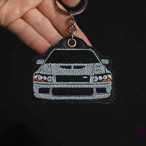 Może przedstawiać: Brelok do kluczyków samochodowych w kolorze srebrnym i czarnym z haftem przedstawiającym widok z przodu Mitsubishi Lancer Evolution.