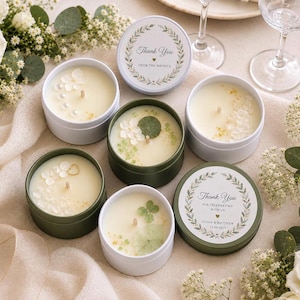 May include: A collection of round metal candle tins in white and olive green, some open to reveal white wax decorated with dried flowers, crystals, and pearls, and others closed with labels reading Thank You from the Mavro's or Olivia & Matthew.