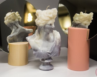 Puede incluir: Dos velas decorativas con forma de bustos de mujer, una de color rosa claro y la otra de color morado claro. Las velas se muestran sobre bases cilíndricas de madera y rosa. Las velas se reflejan en dos espejos redondos.