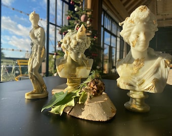 Puede incluir: Tres esculturas de estilo clásico sobre una superficie negra. Las esculturas incluyen una estatua de cuerpo entero y dos bustos, todos en blanco con detalles de pan de oro. El fondo presenta una ventana con vistas y un árbol de Navidad.
